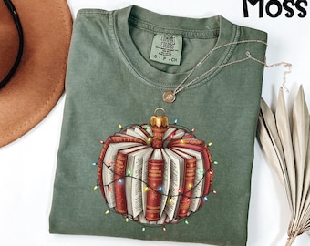 Könnte beinhalten: Ein moosgrünes T-Shirt mit einem Weihnachtsbaumschmuck-Design aus Büchern. Der Schmuck ist mit einer Lichterkette umwickelt. Das Wort "Moss" befindet sich in der oberen rechten Ecke.