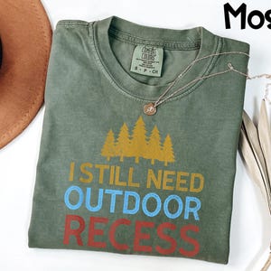 May include: A moss green t-shirt with the text "I STILL NEED OUTDOOR RECESS" in yellow, blue, and red. The shirt features a graphic of yellow pine trees above the text. A gold necklace with a pendant is draped across the shirt.