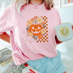 Puede incluir: Camiseta rosa claro con un dise&ntilde;o de calabaza retro, rodeada de flores y un patr&oacute;n de cuadros. La calabaza es naranja y las flores son amarillas y naranjas. Se sostiene una taza blanca con una carita sonriente.