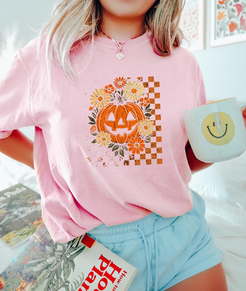 Puede incluir: Camiseta rosa claro con un dise&ntilde;o de calabaza retro, rodeada de flores y un patr&oacute;n de cuadros. La calabaza es naranja y las flores son amarillas y naranjas. Se sostiene una taza blanca con una carita sonriente.