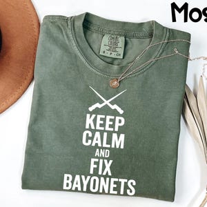 May include: A moss green t-shirt with the text "KEEP CALM AND FIX BAYONETS" in white. The shirt features a crew neck and short sleeves. A gold necklace with a pendant is draped across the shirt. A brown hat and dried plant are in the background.