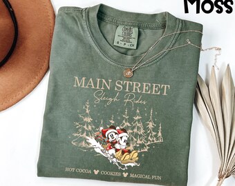 May include: A moss green t-shirt with the text "MAIN STREET Sleigh Rides" and a graphic of Mickey and Minnie Mouse on a sled. The shirt also features the phrases "HOT COCOA", "COOKIES", and "MAGICAL FUN". A gold necklace is draped across the shirt.