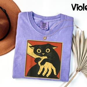May include: A violet-colored t-shirt featuring a graphic of a black cat being held by a hand, set against a red background. The cat has large yellow eyes. The word "Violet" is written in black script in the upper right corner.