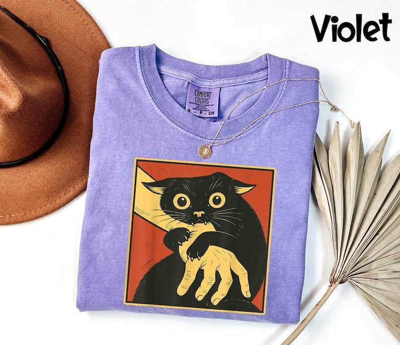 May include: A violet-colored t-shirt featuring a graphic of a black cat being held by a hand, set against a red background. The cat has large yellow eyes. The word "Violet" is written in black script in the upper right corner.