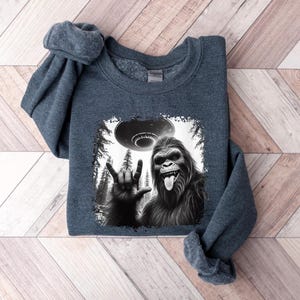 May include: A dark gray sweatshirt with a black and white graphic print of a Bigfoot giving the peace sign with a UFO in the background.