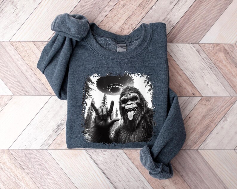 May include: A dark gray sweatshirt with a black and white graphic print of a Bigfoot giving the peace sign with a UFO in the background.