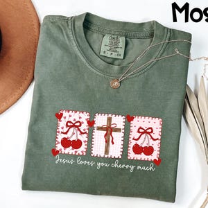 May include: Moss green t-shirt with a graphic featuring cherries, a cross, and the text "Jesus loves you cherry much." The design includes red hearts and bows. A gold necklace is draped across the shirt.