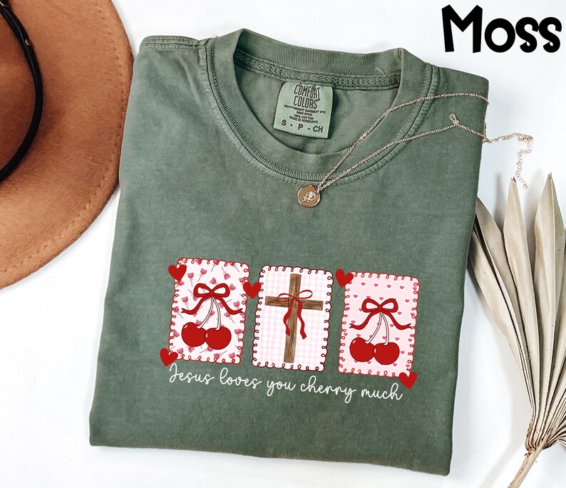 May include: Moss green t-shirt with a graphic featuring cherries, a cross, and the text "Jesus loves you cherry much." The design includes red hearts and bows. A gold necklace is draped across the shirt.