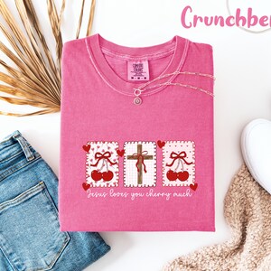 May include: A pink t-shirt with a graphic of cherries, a cross, and the text "Jesus loves you cherry much." The shirt is paired with blue jeans and white sneakers. The word "Crunchberry" is written in pink script.