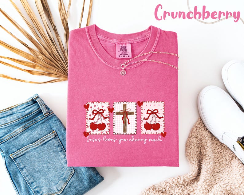 May include: A pink t-shirt with a graphic of cherries, a cross, and the text "Jesus loves you cherry much." The shirt is paired with blue jeans and white sneakers. The word "Crunchberry" is written in pink script.