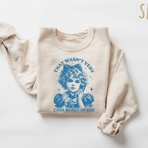 Puede incluir: Una sudadera beige con un gr&aacute;fico azul de una ni&ntilde;a al tel&eacute;fono. El gr&aacute;fico dice "That wasn't very cash money of you".