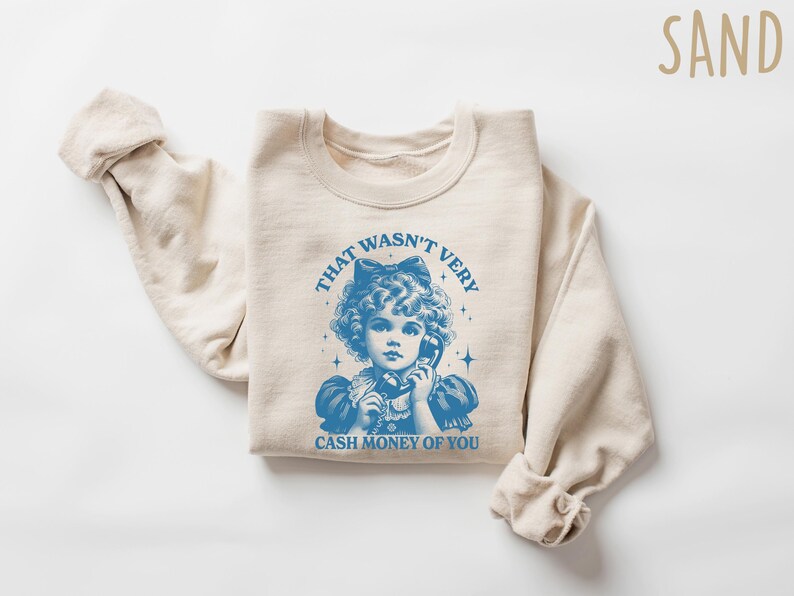 Puede incluir: Una sudadera beige con un gr&aacute;fico azul de una ni&ntilde;a al tel&eacute;fono. El gr&aacute;fico dice "That wasn't very cash money of you".