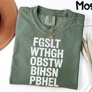 May include: A moss green Comfort Colors t-shirt with white block letters and a gold necklace. The text on the shirt reads "FGSLT WTHGH OBSTW BIHSN PBHEL". The shirt is size S-P-CH.