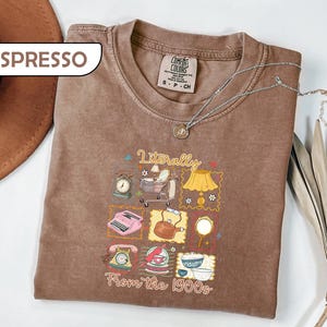 May include: An espresso-colored t-shirt with a graphic print featuring the text "Literally From the 1900s" and various vintage illustrations including a typewriter, rotary phone, tea kettle, lamp, baby carriage, and kitchenware items.