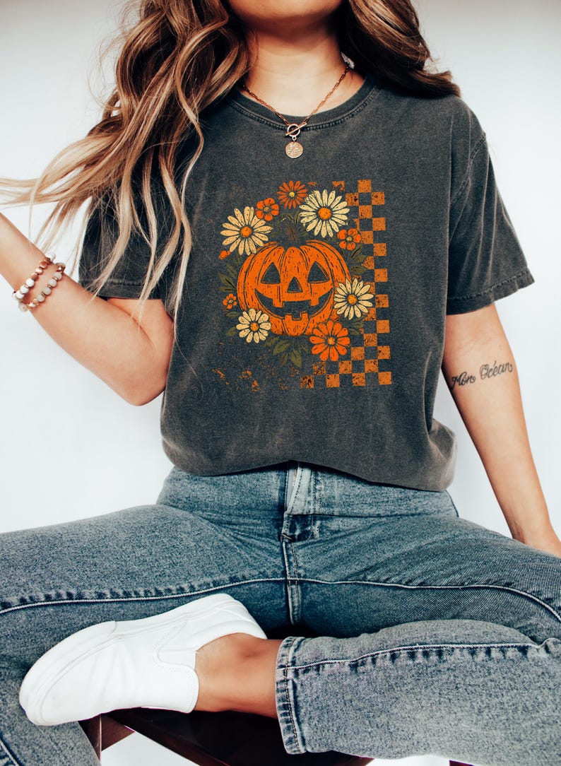 Puede incluir: Camiseta gris oscuro con una calabaza sonriente rodeada de flores naranjas y amarillas. Un patr&oacute;n de cuadros est&aacute; en el lado derecho de la calabaza. La camiseta se usa con jeans azules y zapatos blancos.