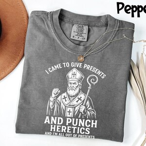 May include: A grey t-shirt featuring the text "I CAME TO GIVE PRESENTS AND PUNCH HERETICS AND I'M ALL OUT OF PRESENTS" and a graphic of a religious figure. The shirt has a crew neck and short sleeves. A gold necklace is draped across the shirt.