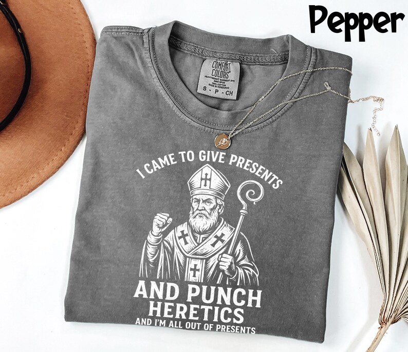 May include: A grey t-shirt featuring the text "I CAME TO GIVE PRESENTS AND PUNCH HERETICS AND I'M ALL OUT OF PRESENTS" and a graphic of a religious figure. The shirt has a crew neck and short sleeves. A gold necklace is draped across the shirt.