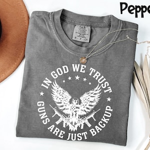 May include: Pepper-colored t-shirt with a crew neck and short sleeves. The shirt features a white graphic of an eagle, guns, and the text "IN GOD WE TRUST, GUNS ARE JUST BACKUP". A gold necklace with a pendant is on the shirt.