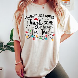Puede incluir: Camiseta color crema con el texto "Probably Just Gonna Jingle Some of the Way, I'm Tired." El dise&ntilde;o incluye un gorro de Pap&aacute; Noel, estrellas y una guirnalda de luces de colores. Prenda casual y festiva.