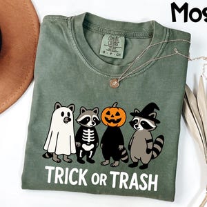 May include: Moss green t-shirt with a Halloween-themed graphic. The graphic features four cartoon raccoons dressed as a ghost, skeleton, jack-o'-lantern, and witch. The text "TRICK OR TRASH" is printed in white below the characters. A gold necklace is visible.