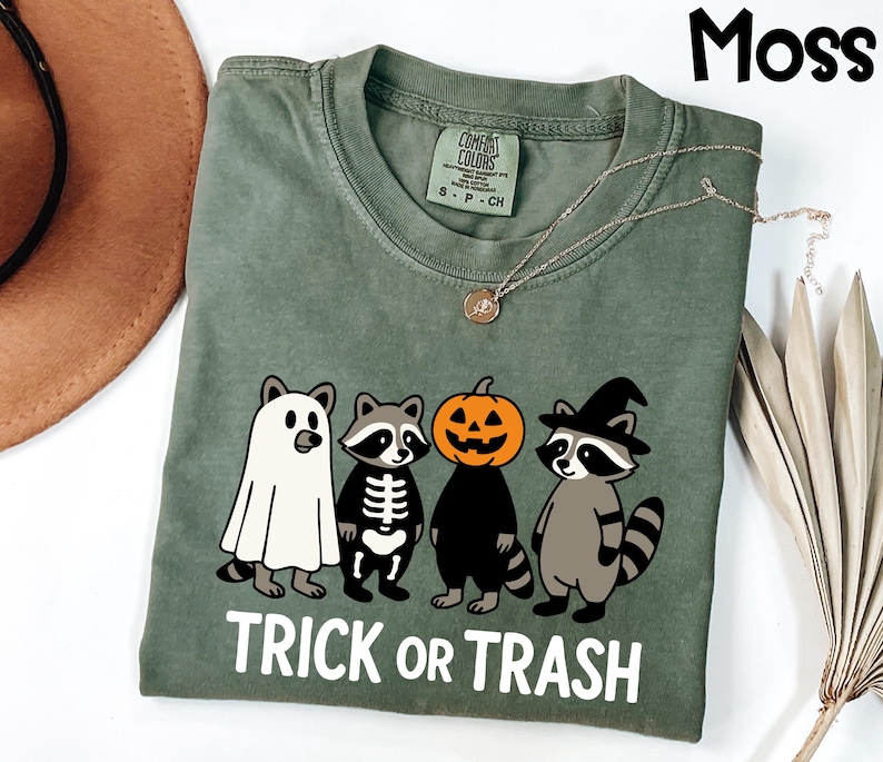 May include: Moss green t-shirt with a Halloween-themed graphic. The graphic features four cartoon raccoons dressed as a ghost, skeleton, jack-o'-lantern, and witch. The text "TRICK OR TRASH" is printed in white below the characters. A gold necklace is visible.