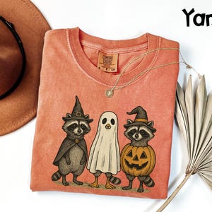 May include: A coral-colored t-shirt featuring a Halloween-themed graphic. The graphic depicts three cartoon characters: two raccoons in witch hats and a ghost. The raccoon on the right is inside a carved pumpkin. The shirt has a crew neck and a Comfort Colors tag.