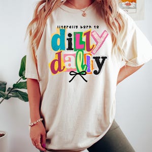以下が含まれることがあります： 「literally born to dilly dally」というフレーズがカラフルで遊び心のある文字でプリントされた、クリーム色のTシャツ。テキストは青、ピンク、緑、黒など様々な色で表現されています。テキストの下には黒いリボンのグラフィック。カジュアルで快適なスタイルです。