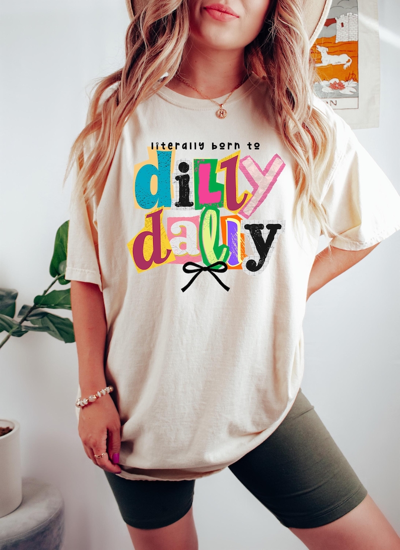 以下が含まれることがあります： 「literally born to dilly dally」というフレーズがカラフルで遊び心のある文字でプリントされた、クリーム色のTシャツ。テキストは青、ピンク、緑、黒など様々な色で表現されています。テキストの下には黒いリボンのグラフィック。カジュアルで快適なスタイルです。