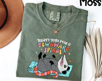May include: Moss green t-shirt with the text "Today's yoga pose is DOWNWARD SPIRAL" and a cartoon opossum on a yoga mat. The opossum is gray with a pink and orange tail. A gold necklace is draped on the shirt.