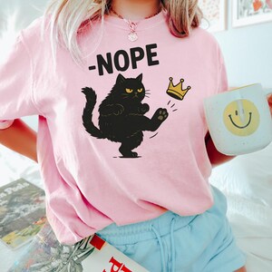 Op de afbeelding: Roze sweatshirt met het woord "-NOPE" boven een zwarte kat die tegen een gouden kroon trapt. De kat heeft een boze uitdrukking. Een witte mok met een gele smiley wordt vastgehouden.