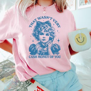 Puede incluir: Camiseta rosa con una ilustraci&oacute;n vintage de una ni&ntilde;a al tel&eacute;fono. El texto en la camiseta dice "That wasn't very cash money of you."