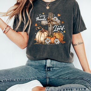 Puede incluir: Camiseta gris oscuro con un dise&ntilde;o gr&aacute;fico que presenta una cruz de madera, calabazas y margaritas. El texto dice "Give thanks to the Lord". El dise&ntilde;o incorpora colores y elementos oto&ntilde;ales, adecuados para temas de oto&ntilde;o o religiosos.