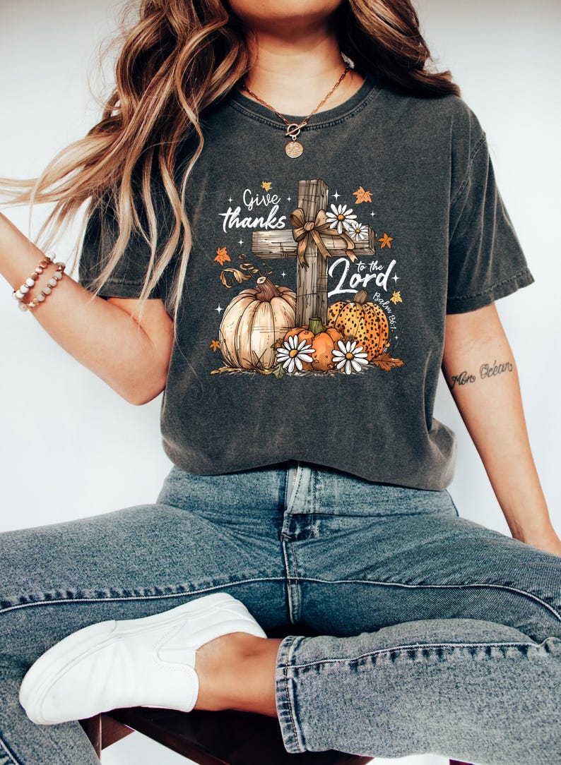 Puede incluir: Camiseta gris oscuro con un dise&ntilde;o gr&aacute;fico que presenta una cruz de madera, calabazas y margaritas. El texto dice "Give thanks to the Lord". El dise&ntilde;o incorpora colores y elementos oto&ntilde;ales, adecuados para temas de oto&ntilde;o o religiosos.