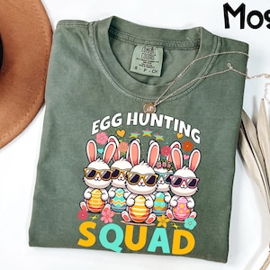 May include: Moss green t-shirt featuring a graphic of five cartoon bunnies wearing sunglasses and holding Easter eggs. The text "EGG HUNTING SQUAD" is printed above and below the bunnies in white and yellow. The shirt has a crew neck and short sleeves.