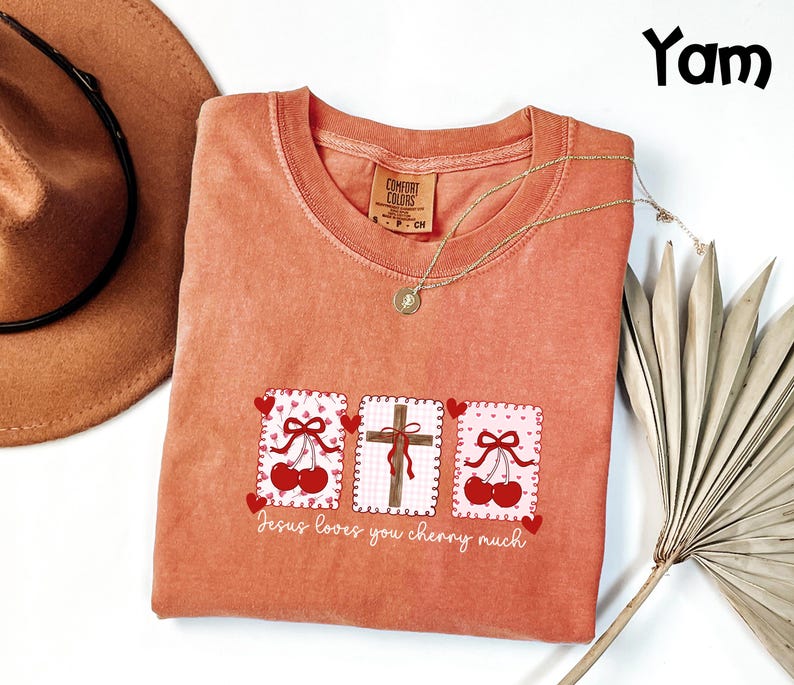 May include: A coral-colored crew neck t-shirt with a graphic design featuring three square patches. The patches display a cross, cherries, and hearts. The text "Jesus loves you cherry much" is printed below the patches. A gold necklace and a brown hat are also visible.