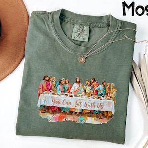 May include: Moss green t-shirt with a crew neck and short sleeves. The shirt features a colorful graphic of a religious scene with the text "You Can Sit With Us" and "Romans 15:7". A silver necklace with a pendant is on the shirt.