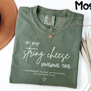 May include: A moss green t-shirt with the text "in my string cheese mama era" in white script. A gold necklace with a pendant rests on the shirt. The shirt has a Comfort Colors tag.