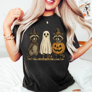 May include: Black t-shirt featuring a Halloween-themed graphic. The design includes three cartoon characters: a raccoon in a witch hat and cape, a ghost, and a raccoon inside a jack-o'-lantern. The artwork has a vintage, hand-drawn style.