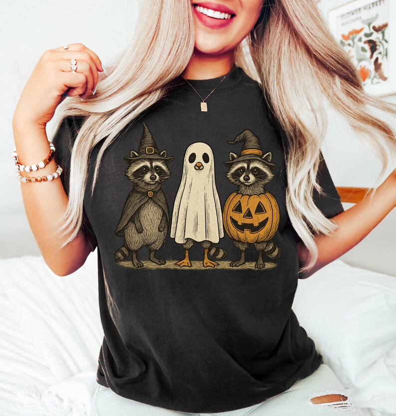 May include: Black t-shirt featuring a Halloween-themed graphic. The design includes three cartoon characters: a raccoon in a witch hat and cape, a ghost, and a raccoon inside a jack-o'-lantern. The artwork has a vintage, hand-drawn style.