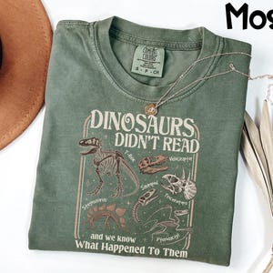May include: A moss green t-shirt with the text "DINOSAURS DIDN'T READ" and dinosaur illustrations. The shirt features a crew neck and short sleeves. A gold necklace with a pendant is draped across the shirt.