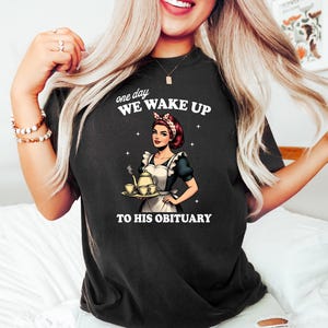 May include: Black t-shirt with a graphic of a woman in a maid outfit holding a teapot and teacups. The text on the shirt reads "one day we wake up to his obituary."