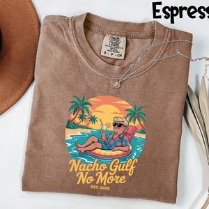 May include: Espresso-colored t-shirt with a graphic of a man relaxing in a pool float with a tropical background. The graphic includes the text "Nacho Gulf No More" and "EST. 2025". The shirt has a crew neck and short sleeves.