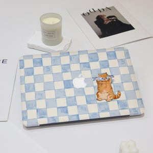 Könnte beinhalten: Laptop-Hülle mit blau-weißem Karomuster und einer orangefarbenen Cartoon-Katze, die zwei Fische hält. Das Apple-Logo ist sichtbar. Eine brennende Kerze und eine Zeitschrift befinden sich im Hintergrund.