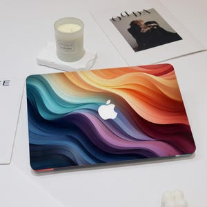 May include: A closed laptop featuring a vibrant, multi-colored abstract wave pattern on its lid. The design consists of layered, flowing curves in shades of deep blue, purple, teal, orange, and yellow. The silver Apple logo is centered on the lid.