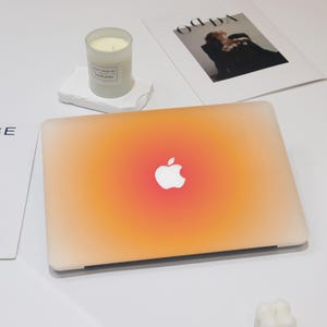 Könnte beinhalten: Ein Laptop mit einem Farbverlauf in Orange und Weiß. Das Apple-Logo ist in der Mitte sichtbar. Eine brennende Kerze in einem Glas steht auf einem weißen Stein, mit einer Zeitschrift im Hintergrund.