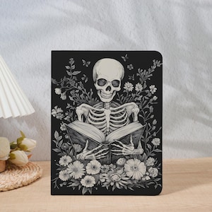 Op de afbeelding: Zwart notitieboek met een grijswaarden illustratie van een skelet dat een boek leest, omringd door bloemen en bladeren. De afbeelding is zeer gedetailleerd. Het notitieboek heeft een matte afwerking en een eigentijds ontwerp. Thema: gothic en botanisch.