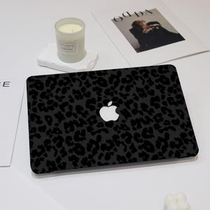May include: A black laptop with a leopard print design and a white Apple logo. A lit candle in a glass jar and a magazine are also on the white surface.