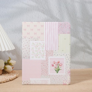 May include: A floral-themed notebook with a patchwork design. The cover features various patterns, including pink bows, stripes, and floral prints. A square image of pink tulips is centered on the cover. The notebook is on a wooden surface.