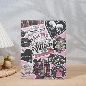 May include: A pink notebook with a collage of stickers. The stickers include a heart with barbed wire, a coffin, skulls, roses, and the text "Falling for the Villain" and "Don't Touch Me".
