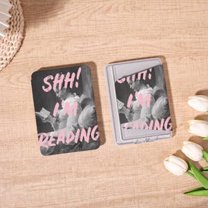 Könnte beinhalten: Zwei rechteckige Kartenetuis. Eines ist offen, das andere geschlossen. Beide zeigen ein Schwarz-Weiß-Bild einer lesenden Person mit rosa Text "SHH! I AM READING".